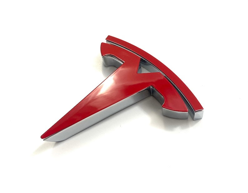 K&ouml;nnte beinhalten: Rotes und silbernes Tesla-Logo-Emblem. Das Logo ist ein stilisiertes 'T' mit einer abgerundeten Oberseite und einer spitzen Unterseite.