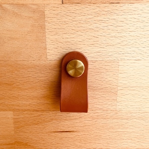 Può includere: Un tirante per cassetti in pelle marrone con un accento metallico circolare color ottone. Il tirante ha una forma rettangolare semplice con bordi arrotondati ed è progettato per una facile presa. L'hardware è di un tono caldo e neutro.