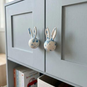May include: Two ceramic cabinet knobs shaped like rabbit heads with long ears. The knobs are white with a blue speckled pattern and are mounted on the two doors of a light gray cabinet. The rabbit ears are textured with visible carved lines.