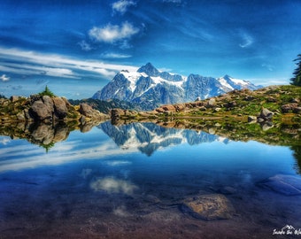 May include: A mountain lake with a reflection of the snow-capped mountains and clouds in the water. The lake is surrounded by rocks and green vegetation.