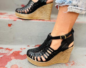 Puede incluir: Sandalias de cuña de cuero negro con un diseño tejido y una suela gruesa de yute de color natural. Las sandalias tienen una correa de tobillo con cierre de hebilla.