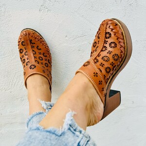 May include: Tan leather clogs featuring a floral cut-out pattern. These clogs have a wooden platform sole and a block heel. The shoes are worn with light blue denim jeans.