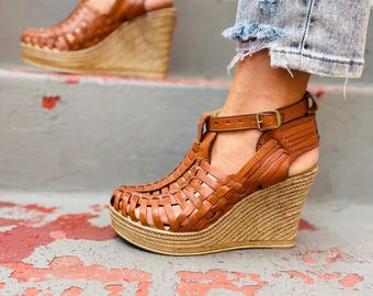 Puede incluir: Sandalias de cuña de cuero marrón con un diseño tejido y una suela gruesa de yute de color natural.