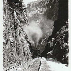 Op de afbeelding: Zwart-witfoto van een trein die door een smalle kloof rijdt. De trein stoot een grote wolk rook uit. De kloofwanden zijn steil en rotsachtig. De afbeelding is gelabeld "Royal Gorge, Canon City, Colo. 1913."