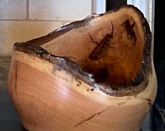 Op de afbeelding: Een handgemaakte houten kom met een natuurlijke rand. De kom heeft een lichtbruin interieur en een donkerbruine, getextureerde schorsrand. De houtnerf is zichtbaar en draagt bij aan de rustieke esthetiek. De kom wordt waarschijnlijk voor decoratieve doeleinden gebruikt.