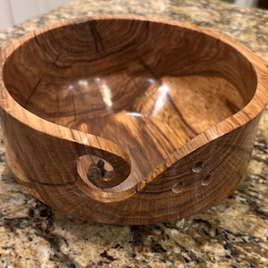 May include: A polished wooden yarn bowl featuring a natural brown wood grain pattern, a smooth interior, and a distinctive spiral cutout on the side for feeding yarn, accompanied by three small circular holes drilled into the side wall.
