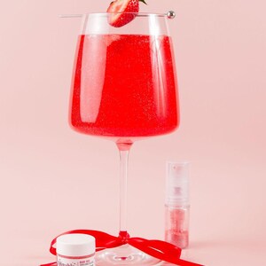 May include: A tall, clear glass filled with a vibrant red liquid, garnished with a strawberry. A small jar and spray bottle of glitter are next to the glass. A red ribbon is tied around the base of the glass. The jar has the text "Bubble Dust".