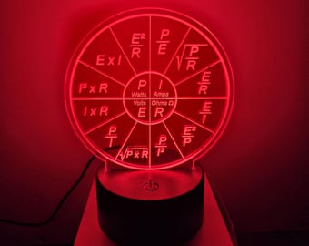 May include: A red-lit, circular LED lamp featuring electrical formulas. The lamp displays equations for Watts, Volts, Amps, and Ohms. The lamp sits on a black base with a silver-colored platform.