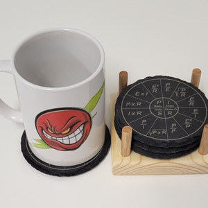 May include: A white ceramic mug with a red and green cartoon face design. The mug is sitting on a black coaster. Behind the mug is a set of four black coasters with white text and formulas related to electricity. The coasters are on a wooden stand.