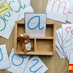 Op de afbeelding: Een set printbare cursieve alfabetkaarten met een blauwe cursieve letter 'a' op een witte kaart. De kaarten zitten in een houten doos met een klein knuffel eekhoorn. De tekst 'PDF imprimible' staat op een geel label.