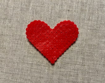 Può includere: Un mosaico di perline a forma di cuore rosso su uno sfondo di tessuto grigio chiaro.