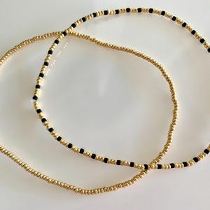 Op de afbeelding: Twee gouden armbandjes met kralen, een met zwarte kralen ertussen.