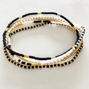 Può includere: Un set di cinque braccialetti con perline nere, bianche e dorate. I braccialetti sono sovrapposti e hanno un design minimalista.