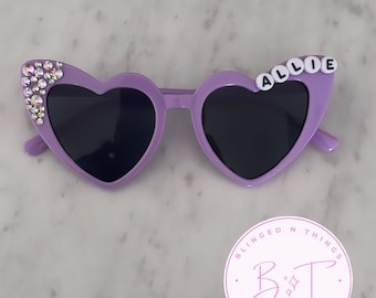 May include: Purple heart-shaped sunglasses with rhinestones on one side and the name "ALLIE" spelled out with white beads on the other side. The sunglasses are on a white marble surface with a pink circle sticker that says "BLINGED N THINGS" with a stylized "B&T" logo.