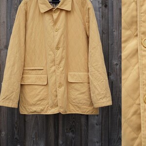 May include: A tan quilted jacket with a button-up front, two front pockets, and a collared neckline. The jacket is hanging on a black hanger against a wooden background. The jacket's diamond pattern is visible.