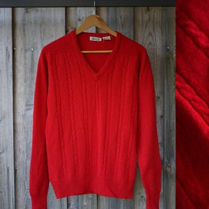 Puede incluir: Un suéter rojo brillante con cuello en V y detalles de punto trenzado. El suéter se muestra en una percha de madera sobre un fondo de madera desgastada. Un primer plano muestra la textura del tejido.