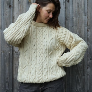 Könnte beinhalten: Ein cremeweißer, langärmeliger Zopfmuster-Pullover mit Rundhalsausschnitt und komplexen vertikalen Flechtmustern. Das Kleidungsstück ist aus einem dicken, strukturierten Garn gefertigt und zeigt ein klassisches Grobstrickdesign.