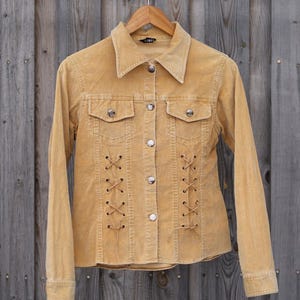 May include: Mustard-colored corduroy jacket with a collared neckline, two chest pockets, and silver buttons. The front features decorative criss-cross lacing. The jacket is hanging on a wooden hanger against a wooden background.