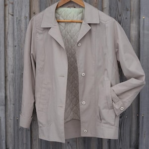 May include: A beige jacket with a quilted lining and a button closure. The jacket is hanging on a wooden hanger against a wood panel background.