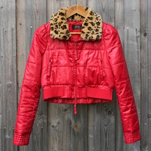 May include: A bright red cropped puffer jacket featuring a faux fur collar with a brown and black leopard print pattern. The jacket has a front zip closure, long sleeves with ribbed cuffs, and a wide ribbed hem. The label reads VeeKo.