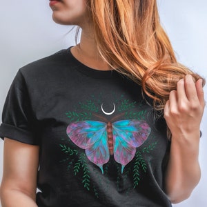 Op de afbeelding: Een zwart T-shirt met een kleurrijke vlinderprint en een halve maan. De vlinder heeft roze, blauwe en paarse vleugels met groen gebladerte eromheen. Het shirt wordt gedragen door iemand die een witte mok vasthoudt met de tekst "You Got This" in kleurrijke letters.