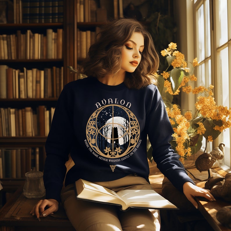 May include: A navy blue sweatshirt with a gold design featuring a full moon, trees, and the text "Avalon" and "Where the name Arthur whispers gently on the breeze".