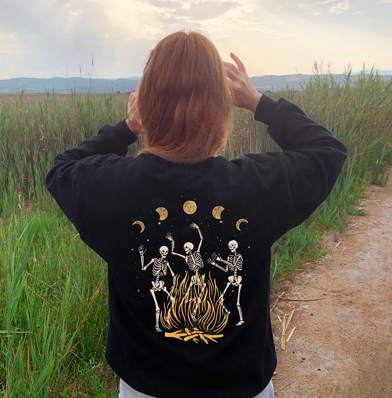 May include: A black sweatshirt with a graphic print of three skeletons dancing around a fire. The skeletons are white and the fire is orange. There are five crescent moons in a row above the skeletons. The moons are yellow.