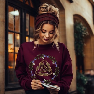 May include: A maroon sweatshirt with a circular design featuring three brown rabbits in a triangle formation surrounded by green leaves and purple flowers.