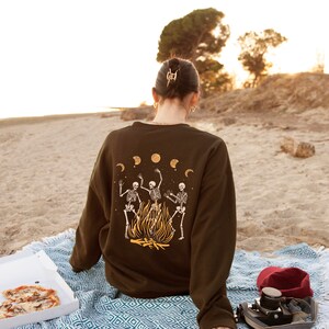 May include: A person wearing a brown sweatshirt with a graphic of three skeletons dancing around a fire. The sweatshirt has a moon and stars design. The person is sitting on a blue and white blanket on a sandy beach.