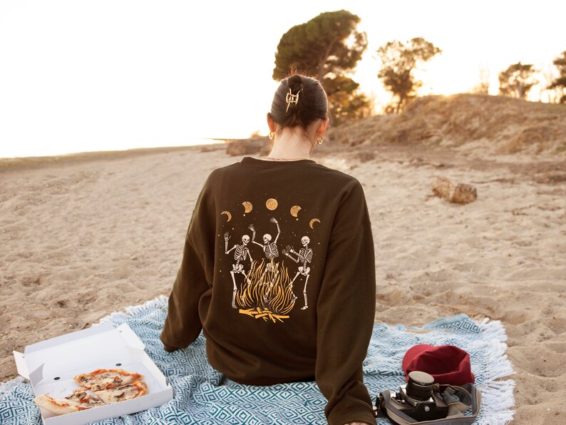 May include: A person wearing a brown sweatshirt with a graphic of three skeletons dancing around a fire. The sweatshirt has a moon and stars design. The person is sitting on a blue and white blanket on a sandy beach.