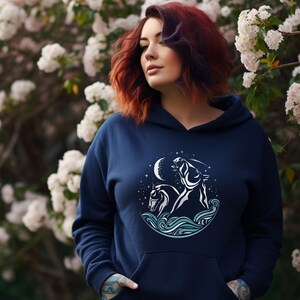 Puede incluir: Una sudadera con capucha azul marino con un gr&aacute;fico blanco de un caballo y un jinete en un c&iacute;rculo con una luna y estrellas. El gr&aacute;fico est&aacute; sobre un fondo de olas azul y blanco.