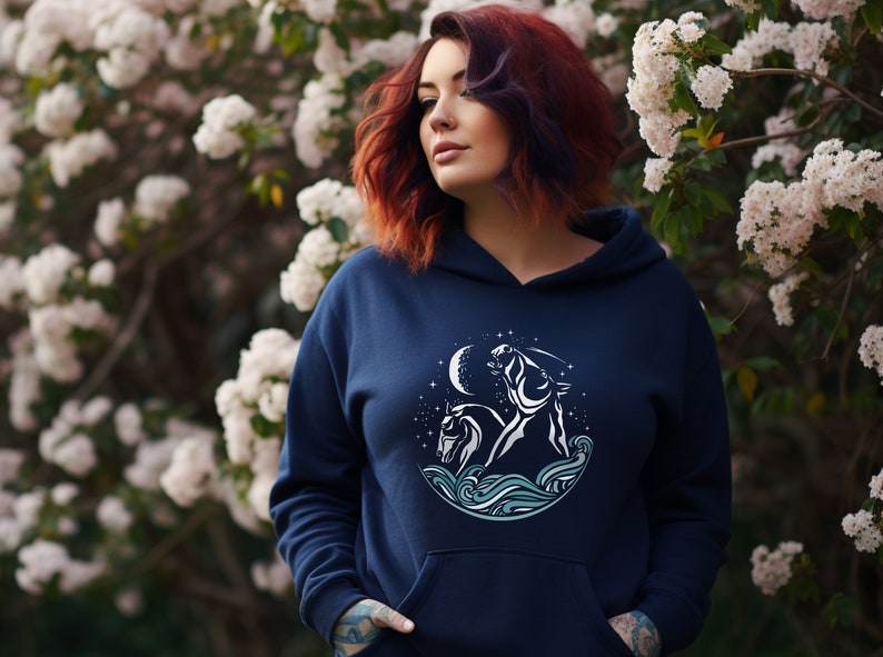 Puede incluir: Una sudadera con capucha azul marino con un gr&aacute;fico blanco de un caballo y un jinete en un c&iacute;rculo con una luna y estrellas. El gr&aacute;fico est&aacute; sobre un fondo de olas azul y blanco.