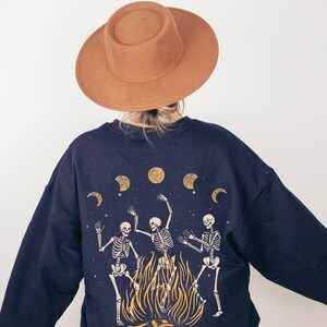 May include: A dark blue sweatshirt with a graphic print of three skeletons dancing around a bonfire with moons and stars in the background. The sweatshirt has long sleeves and a relaxed fit.