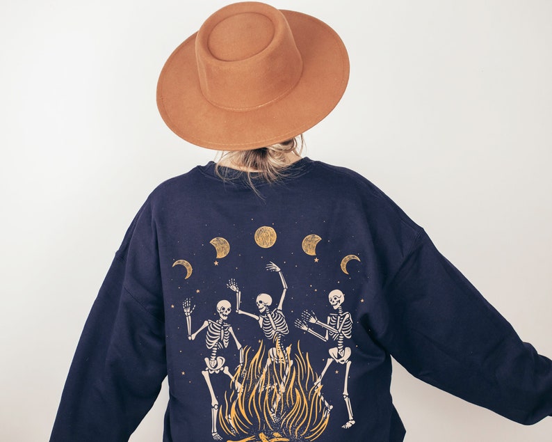 May include: A dark blue sweatshirt with a graphic print of three skeletons dancing around a bonfire with moons and stars in the background. The sweatshirt has long sleeves and a relaxed fit.