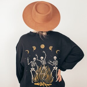 May include: Black sweatshirt with a graphic of three skeletons dancing around a bonfire with moons in the background. The sweatshirt has a brown felt fedora on top.