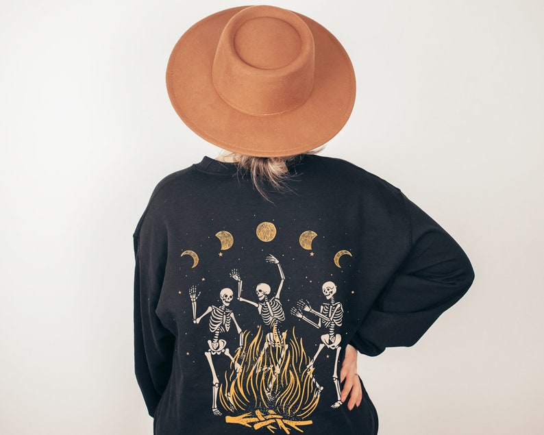May include: Black sweatshirt with a graphic of three skeletons dancing around a bonfire with moons in the background. The sweatshirt has a brown felt fedora on top.
