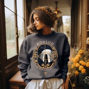 May include: A dark gray sweatshirt with a gold graphic design featuring a full moon, trees, and the text "Avalon" and "Where the name Arthur whispers gently on the breeze".
