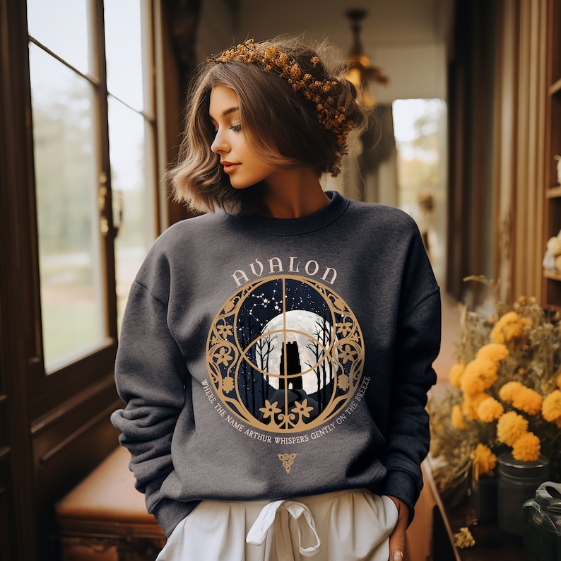 May include: A dark gray sweatshirt with a gold graphic design featuring a full moon, trees, and the text "Avalon" and "Where the name Arthur whispers gently on the breeze".