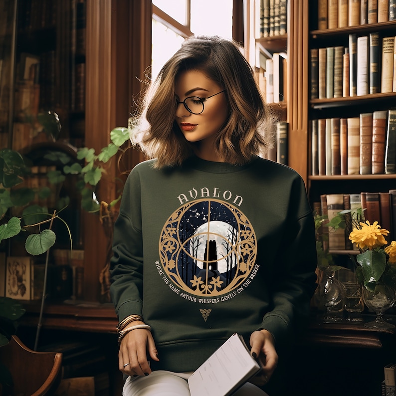 May include: A green sweatshirt with a gold graphic of a moon, trees, and the words "Avalon, Where the Name Arthur Whispers Gently on the Breeze".