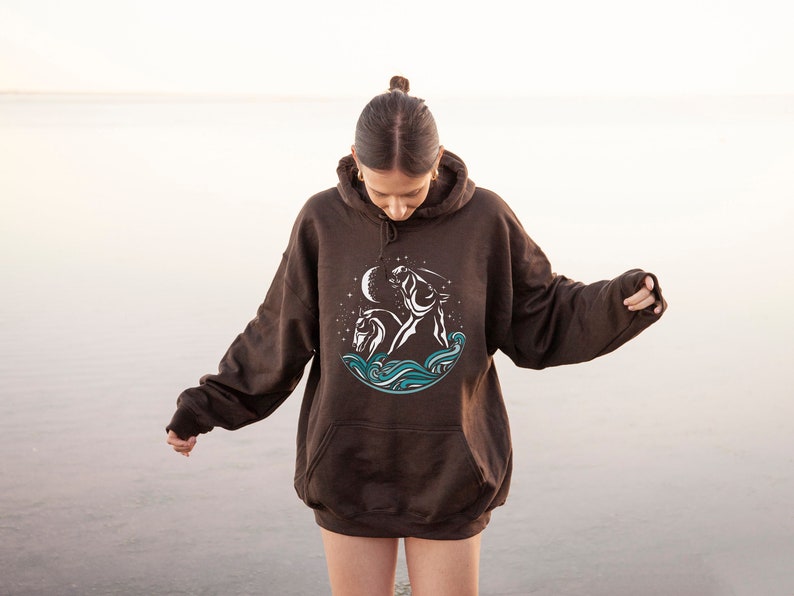 Puede incluir: Una sudadera con capucha marr&oacute;n con un dise&ntilde;o gr&aacute;fico blanco de un caballo y un jinete en el oc&eacute;ano con una luna creciente y estrellas.