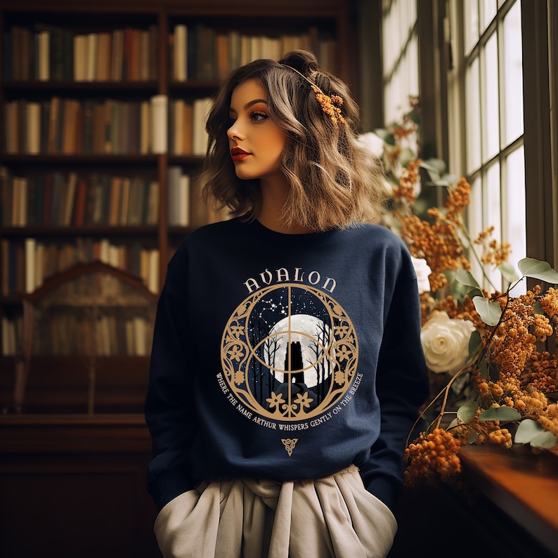 May include: A navy blue sweatshirt with a gold graphic of a moon, trees, and the text "Avalon" and "Where the name Arthur whispers gently on the breeze".