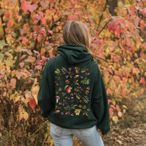May include: A dark green hoodie with a colorful print of mushrooms, leaves, and berries on the back. The hoodie has a drawstring hood and a kangaroo pocket.