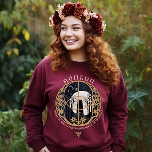May include: A maroon sweatshirt with a gold circular design featuring a moon, trees, and the text "Avalon" and "Where the name Arthur whispers gently on the breeze".
