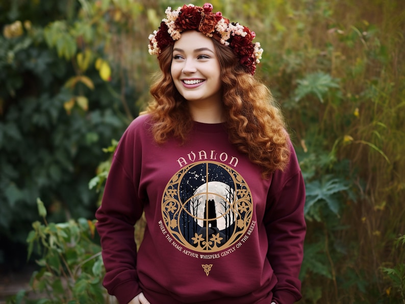 May include: A maroon sweatshirt with a gold circular design featuring a moon, trees, and the text "Avalon" and "Where the name Arthur whispers gently on the breeze".