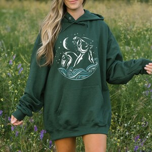 Puede incluir: Una sudadera con capucha verde oscuro con un gr&aacute;fico blanco de dos caballos corriendo a trav&eacute;s de olas y una luna creciente. El gr&aacute;fico est&aacute; rodeado de estrellas.