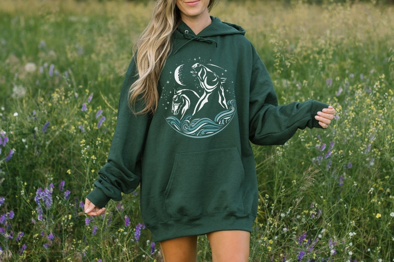 Puede incluir: Una sudadera con capucha verde oscuro con un gr&aacute;fico blanco de dos caballos corriendo a trav&eacute;s de olas y una luna creciente. El gr&aacute;fico est&aacute; rodeado de estrellas.