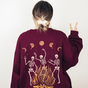 May include: Burgundy sweatshirt with a graphic of three skeletons dancing around a bonfire with moons and stars in the background.