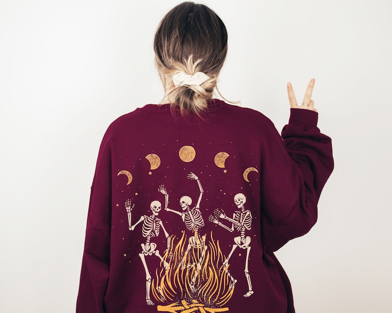 May include: Burgundy sweatshirt with a graphic of three skeletons dancing around a bonfire with moons and stars in the background.