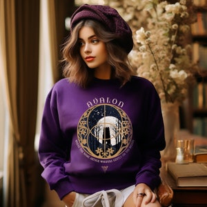 May include: Purple sweatshirt with a gold graphic of a castle in a forest with a full moon and stars. The graphic has the text "Avalon" and "Where the name Arthur whispers gently on the breeze".