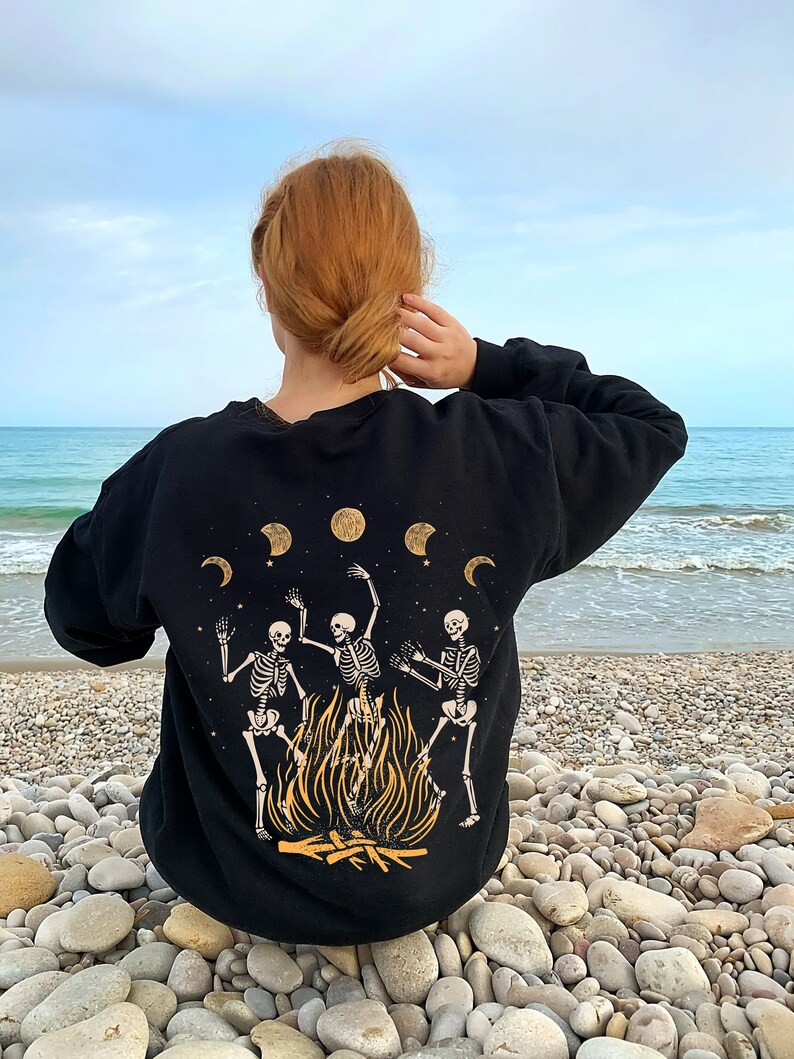 May include: Black sweatshirt with a graphic of three skeletons dancing around a bonfire. The graphic is in gold and yellow. There are five crescent moons in the background.
